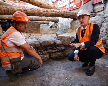 Dos hombres con casco de seguridad y chaleco reflectante están agachados junto a una pared de piedras en una excavación