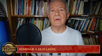 Un hombre situado frente a una biblioteca con un micrófono a un costado y un gráfico con texto en el sector inferior de la pantalla