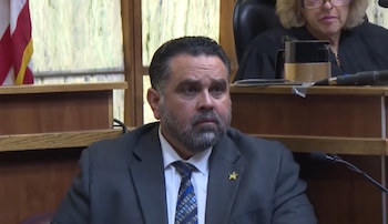 Un hombre con barba, traje gris y corbata testifica sentado en un estrado de madera dentro de una sala de tribunal
