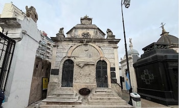 Mausoleo de mármol con techo abovedado, dos puertas de rejas, escaleras, estatuas y otros sepulcros en un cementerio bajo cielo nublado