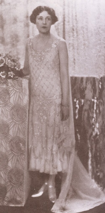 Fotografía en tono sepia de una mujer con vestido bordado de tirantes y velo que apoya su mano sobre una columna decorada