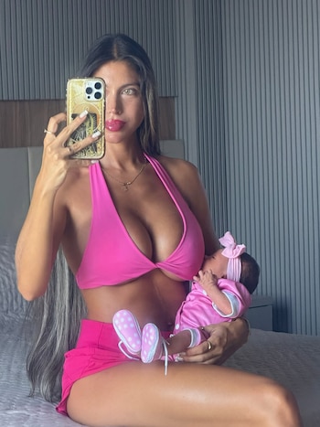 Una mujer vestida de rosa sostiene a un bebé en brazos mientras se toma una fotografía frente a un espejo con un celular