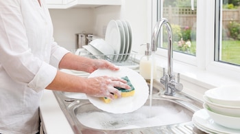 Manos de una persona sostienen una esponja jabonosa y un plato blanco junto a un grifo abierto en el fregadero de una cocina.