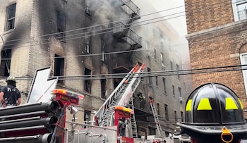 Edificio de ladrillo con fachadas quemadas y humo, camión de bomberos con escalera extendida, varios bomberos y casco en primer plano