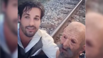 Un hombre de camisa y chaleco posa junto a una persona con una máscara de rostro arrugado y barba gris sobre rieles de tren