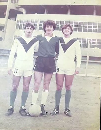 Tres jóvenes futbolistas uniformados, uno con camiseta verde y pantalón corto negro, y dos con camisetas blancas con V verde, posan con un balón en un campo de tierra