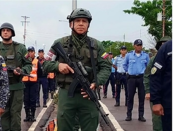 Un militar con casco, uniforme verde oliva y chaleco táctico sostiene un rifle de asalto en primer plano, con otros militares y policías de fondo