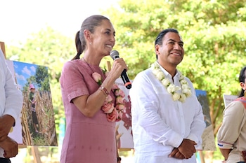Claudia Sheinbaum habla con micrófono y collar de rosas. Eduardo Ramírez sonríe con collar de flores blancas. Fondo borroso con árboles y un panel con imagen