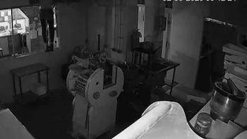 Imagen monocromática de una persona subiendo por una ventana en un cuarto oscuro con maquinaria industrial y mesas en primer plano
