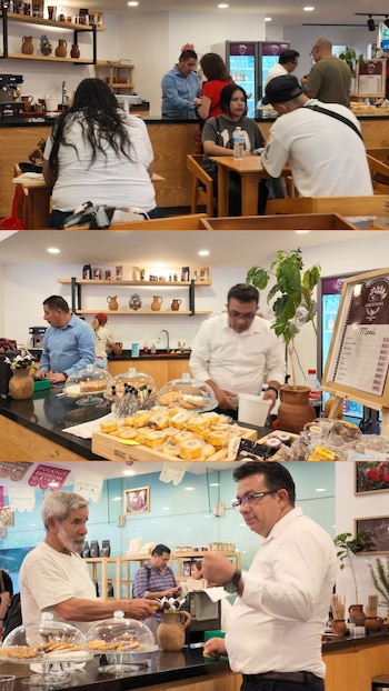 Múltiples personas en una cafetería moderna con mesas de madera, un mostrador exhibiendo pasteles y galletas bajo campanas de cristal, y estanterías con jarrones artesanales