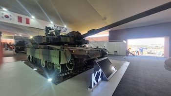 Un tanque de batalla K2 Black Panther de camuflaje, con cañón largo y orugas, se exhibe en un pabellón con banderas de Corea del Sur y Perú, y logotipos de Hyundai y Kia