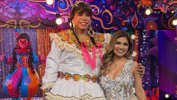 Dos personas posan en un escenario de televisión. Una lleva un vestido blanco bordado, la otra viste un vestido brillante. De fondo, una figura de payaso y luces