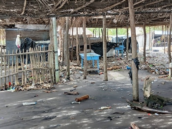 Interior de una estructura abierta tipo choza con techo de palma seca y postes de madera, el suelo oscuro cubierto de abundante basura y escombros, con algunas mesas azules