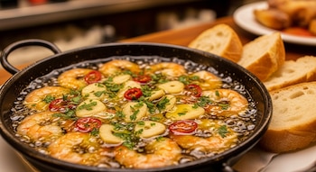 Cazuela negra con gambas al ajillo burbujeantes: camarones rosados, láminas de ajo, guindilla roja y perejil fresco en aceite, con pan rústico al lado.