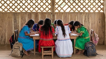 Seis niñas con cabello largo oscuro y vestidos coloridos se sientan en un aula de madera, escribiendo en cuadernos sobre una mesa con lápices. Mochilas en el piso.