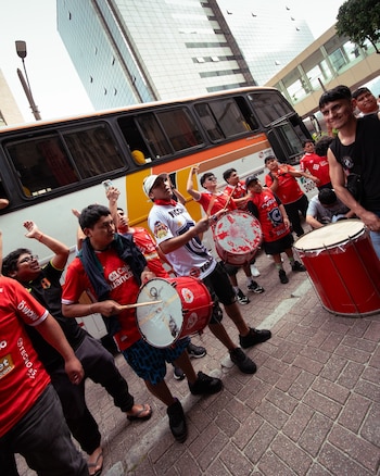 Un grupo de personas con camisetas rojas y blancas toca tambores y aplaude junto a un autobús en una calle pavimentada