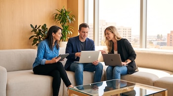 Un hombre y dos mujeres sentados en un sofá, mirando dos computadoras portátiles y una tableta. Detrás hay una pared de madera, plantas y una ventana grande.