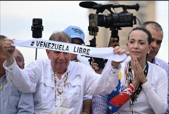 Un hombre y una mujer sostienen una pancarta blanca con el texto '#VENEZUELA LIBRE'. Al fondo, un camarógrafo con una cámara de televisión y un móvil