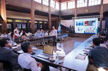 Un orador calvo presenta en un salón de conferencias con techo alto de madera. Asistentes escuchan sentados en mesas mientras una pantalla muestra gráficos