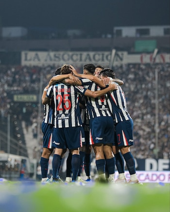 Un grupo de jugadores de fútbol con uniformes de rayas blancas y azules se abrazan en un festejo en el campo. El fondo muestra una grada llena de aficionados