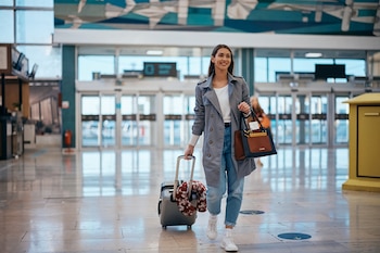 Una mujer joven con abrigo gris camina por un pasillo de aeropuerto, arrastrando una maleta gris con una almohada de viaje y llevando un bolso marrón y negro