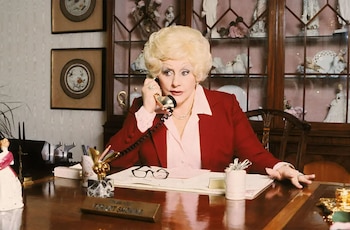 Mary Kay Ash, una mujer rubia con peinado voluminoso, viste una chaqueta roja y camisa rosa, hablando por un teléfono dorado en un escritorio de madera