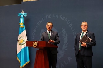 Hombre con gafas y traje hablando en un podio con micrófono y emblema. Otro hombre de barba y gafas sostiene documentos. Bandera de Guatemala a la izquierda