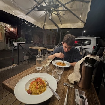 Hombre de camiseta oscura come pasta en plato blanco. Mesa de madera con otro plato de pasta, botellas de agua y vino, copas, cámara de fotos. Terraza exterior de noche