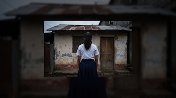 Una joven de espaldas con camisa blanca y falda azul está frente a una vivienda de paredes desgastadas y techo de lámina.
