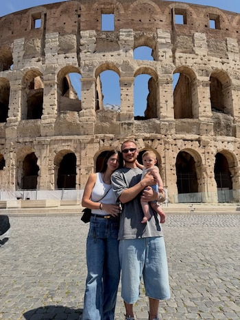 Ailén Cova, Alexis Mac Allister con su hija Alaia en brazos, posando frente al Coliseo de piedra antigua bajo un cielo azul claro
