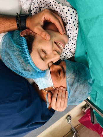 Primer plano de una mujer en gorro quirúrgico y camisón de hospital, sonriendo con ojos cerrados, abrazada por un hombre en uniforme médico y mascarilla