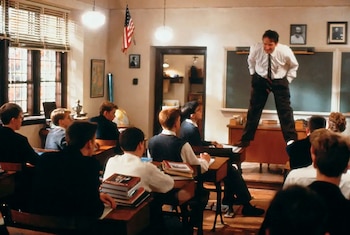 Un hombre de camisa blanca y corbata está de pie sobre un pupitre frente a varios estudiantes sentados en un aula, con una bandera de Estados Unidos en la pared