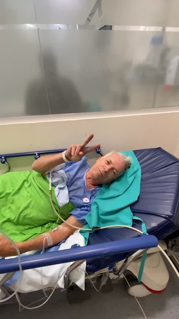 Gerardo Romano en camilla de hospital con bata azul, vía intravenosa en el brazo y haciendo el signo de la paz. Cubierto con sábana verde