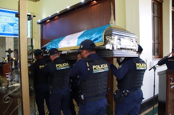Dos agentes de policía en uniforme táctico cargan un ataúd gris adornado. Una fila de policías de uniforme blanco y azul saluda