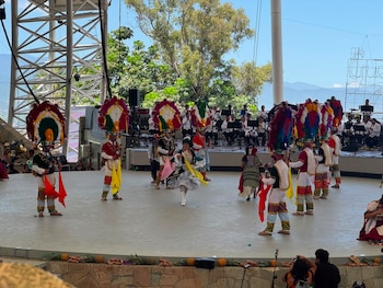 Escenario con once bailarines en trajes típicos, una bailarina de blanco al centro. Detrás, una orquesta. Al fondo, árboles y montañas. Piñas en el borde del escenario