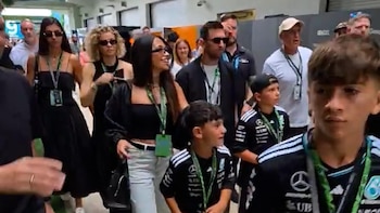Lionel Messi con gafas de sol y su familia, incluyendo Antonela Roccuzzo y sus hijos vestidos con atuendos de Mercedes-Benz, caminan por un área interior