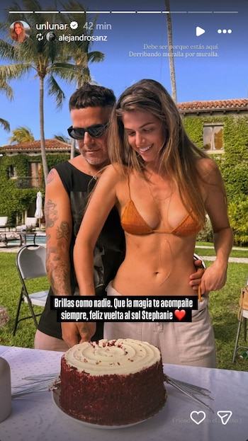 Alejandro Sanz con camiseta negra y gafas de sol abraza a Stephanie Cayo, quien viste bikini top naranja y pantalón claro, con un pastel red velvet en primer plano al aire libre