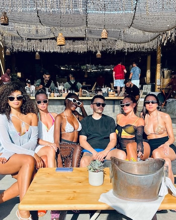 Un grupo de seis personas, cinco mujeres y un hombre, con gafas de sol, sentados en una mesa de madera al aire libre con bebidas