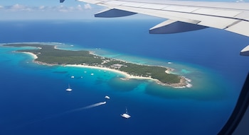Vista desde la ventanilla de un avión con su ala visible, una isla tropical con playas de arena blanca, aguas turquesas, vegetación y pequeñas embarcaciones.