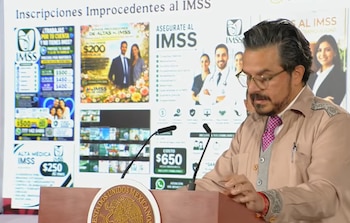 Hombre con barba y lentes habla en podio con escudo de México y micrófonos. Pantalla detrás muestra anuncios fraudulentos de afiliación al IMSS