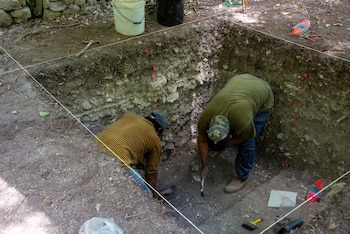 Dos hombres cavan un pozo cuadrado en el suelo. Cuerdas blancas delinean el área superior. Se observan capas de tierra, rocas, marcadores rosados, cubos y herramientas