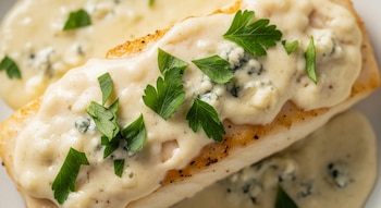 Primer plano de un filet de merluza con piel dorada cubierto por una salsa cremosa de color claro con trozos azules de queso y hojas de perejil fresco.