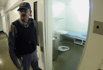 Un oficial de prisión de pie en un pasillo junto a una puerta abierta que muestra el interior de una celda con una cama, un inodoro y un lavamanos