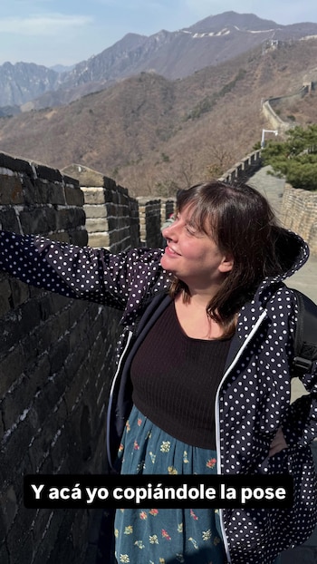 Laila Roth, una mujer de cabello castaño, sonríe de perfil con un abrigo negro de lunares blancos y una falda floral, de pie en la Muralla China con montañas áridas de fondo