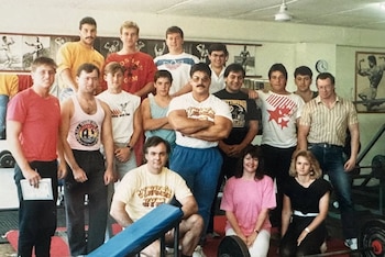 Un grupo mixto de quince personas, en su mayoría hombres, posa en un gimnasio con equipo de levantamiento de pesas y pósteres de culturismo en las paredes