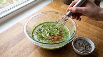 Una mano sostiene un batidor dentro de un recipiente de vidrio con líquido verde y especias, junto a un recipiente pequeño con semillas de chía.