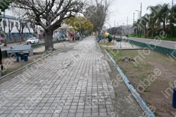 Camino de adoquines, árboles, bancos de hormigón, juegos infantiles, y edificios en el fondo de un parque municipal