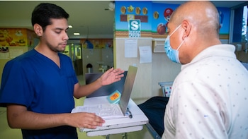 Um jovem com uniforme médico azul parado ao lado de uma cama de hospital, segurando uma pasta e conversando com um idoso mascarado