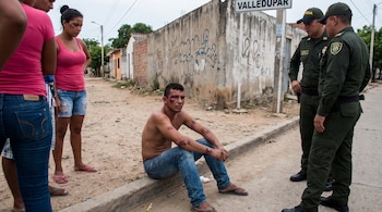 Un hombre sin camisa, con heridas en la cara y brazos, se sienta en la acera. Dos policías están de pie cerca. Un letrero dice "Valledupar".