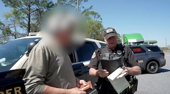 Un oficial de policía con gorra y uniforme negro conversa con un hombre borroso que sostiene una cartera. Un coche de policía y un camión verde se ven detrás